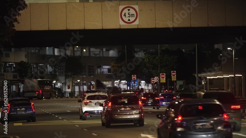 Cars drive along a city street under a bridge at night in Singapore. This scene captures the movement of urban traffic, the city's modern infrastructure, and the nighttime atmosphere.
