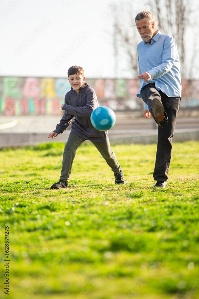 Fototapeta premium Smiling boy and grandfather kicking soccer ball together