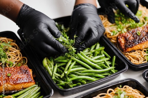 Hands in black gloves arrange fresh food into black plastic containers.  Healthy meal prep.  Focus on vibrant green beans, noodles, and fish