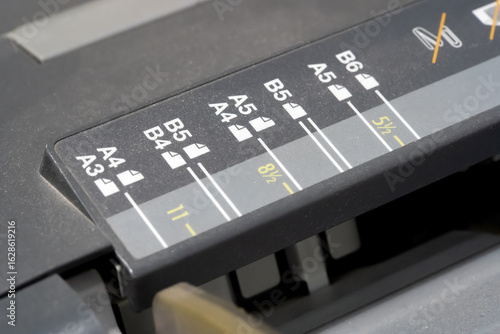Paper size selector panel on office printer or cutter machine showing A3 A4 A5 B4 B5 B6 marks with orientation icons adjustable tray guide scale for 8.5 by 11 inch sheet professional equipment closeup