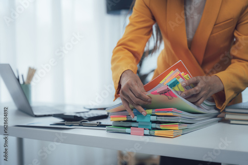 A businesswoman is working with a large pile of paperwork, searching for unfinished documents, information on piles of documents on her desk, and checking financial documents amidst her busy workload.