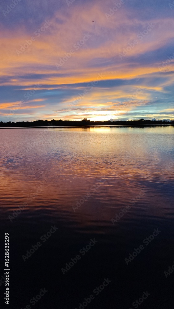 Fototapeta premium Breathtaking sunset over Amazon Jungle River, with vibrant orange and purple clouds reflecting on the calm water surface. A silhouetted forest lines the distant horizon under a dramatic, colorful sky.