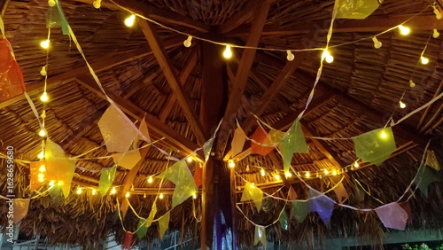 Fotografi Looking up at a rustic thatched roof decorated with colorful festive pennant flags and warm string lights