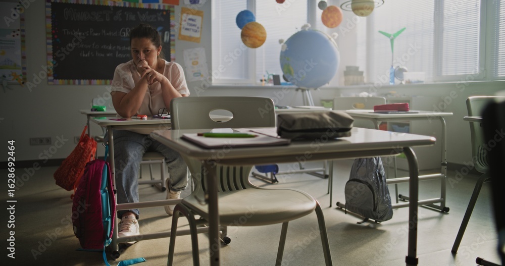 Obraz premium Exhausted Female Teacher Rubbing her Temples, Sitting Alone at Desk in Empty Classroom, Having Rest After Lesson. Woman Feeling Stress, Burnout and Emotional Struggle, Working in Modern Primary School
