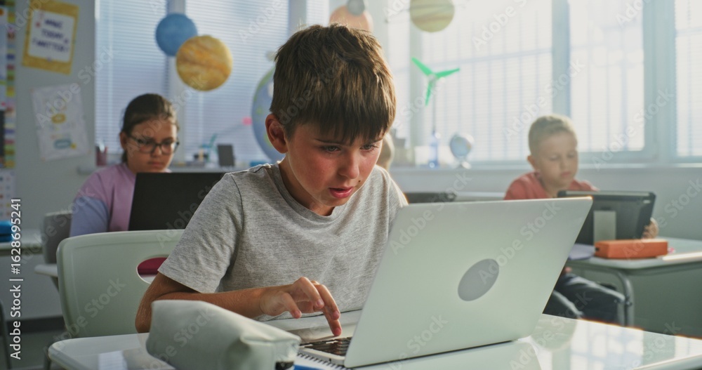 Fototapeta premium Elementary School Boy Sitting at Desk, Using Laptop in Classroom. Team of Smart Diverse Kids Working on Their Laptops and Tablet Computers, Studying Computer Science, Basic Programming and Coding.