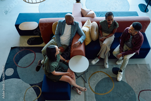 Group of diverse coworkers sitting on colorful sofas having casual conversation in office lounge