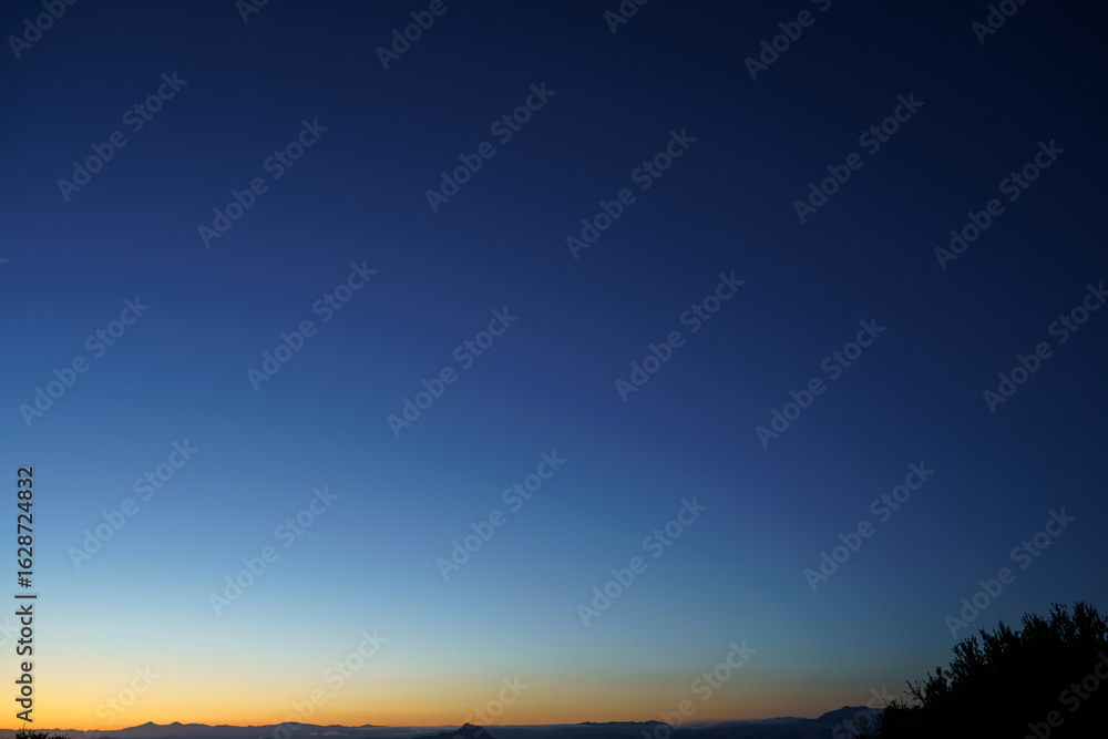 Fototapeta premium Silhouetted treetops and distant mountains lie under a vast, clear sky transitioning from the deep blue of twilight to the warm orange glow of dawn on the horizon.