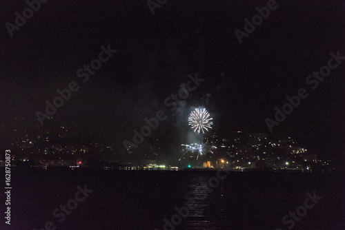 A series of vibrant fireworks light up the night sky over a body of water, with the sparkling lights of a city visible along the distant shore.