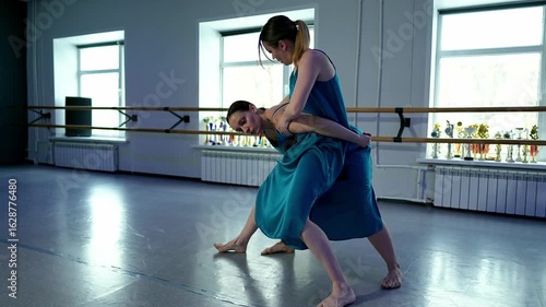 Two Caucasian ballerinas are dancing together and one is lifter her partner filmed in studio at day time. Ladies in blue dresses are performing contemporary sensitive choreography moving to the rhythm