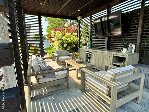 Modern indoor outdoor living entertainment space and patio under a pavilion pergola. poolside equipped with bar and gas fire pit with cascading waterfall. Chair sofa couch and table. Hotel resort feel