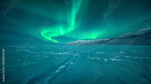 A vast landscape of snowy, icy plains stretches out against a backdrop of snow-capped mountains. Bright green northern lights illuminate the night sky.