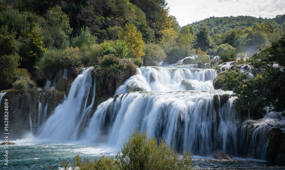Fototapeta premium Peaceful waterfall in a Croatian national park surrounded by lush greenery and clear water. A stunning natural landscape ideal for travel, nature, adventure, and outdoor-themed content.