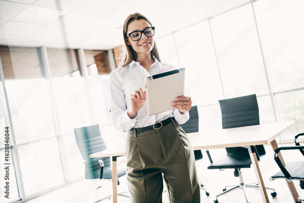Confident young businesswoman working on a tablet in a professional office workspace with large windows and modern furniture. Confident young businesswoman working on a tablet in a professional office workspace with large windows and modern furniture.