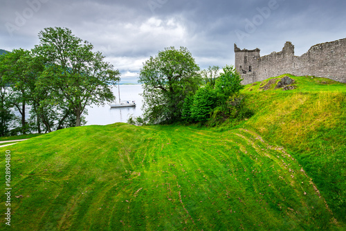 Fotografie The medieval ruins of the hilltop Urquhart Castle above a small boat on Loch Ness lake near Inverness, Scotland, in the Scottish Highlands