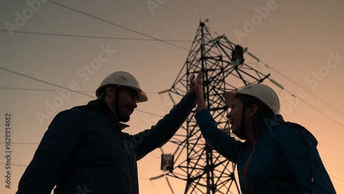 Teamwork, business project on construction of electric communications. Power engineers in protective helmets inspect iron structure of power transmission line. Engineers shake hands. Power engineer