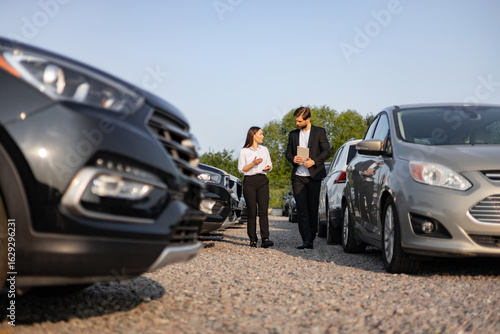 Konstfotografi A male car dealer assists a female customer in a car dealership, potentially during a sale, walking among parked vehicles on a sunny day