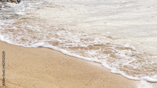 Wallpaper Mural Close-up view of a light sand beach on Aegina Island, Greece. A small wave rolls in from the right with white foam, dissolving gently. Calm coastal scene, partially rocky shore. No people. Torontodigital.ca