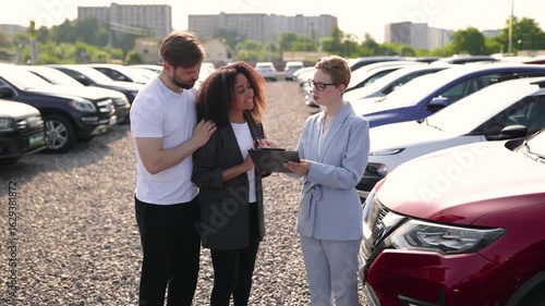 A car salesperson assists a diverse couple with a tablet at a car dealership, surrounded by various vehicles.