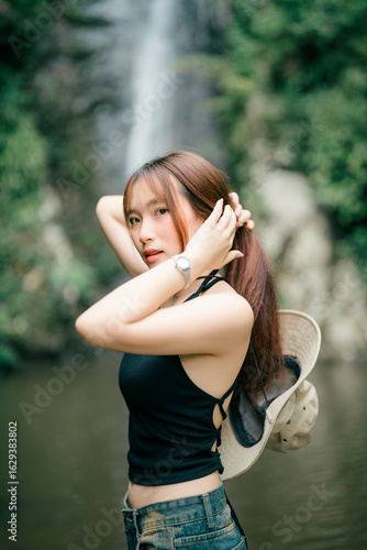 A beautiful young woman poses by a waterfall, tying her hair back as she looks at the camera. A stylish and confident portrait of a traveler enjoying a moment in a lush, scenic paradise.