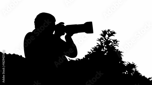 Silhouette of a photographer aiming a telephoto lens from behind foliage against a white background