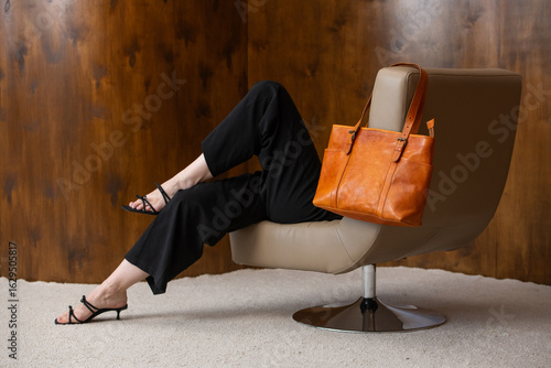 woman with wavy hair seated in a beige chair posing with a rich, vibrant orange leather tote bag. wearing sleeveless black top, trousers that drape gracefully to just above her black heeled sandals.