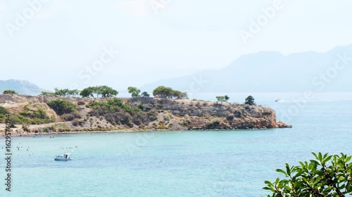 Wallpaper Mural View of a turquoise bay with people swimming near a moored boat, rocky shoreline with dry grass and sparse trees, boat trails on calm water, and hazy hills on the horizon under clear sky Torontodigital.ca