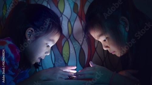 Two Asian child lying in bed using smartphone at night, screen light illuminating face, concept of screen time and digital addiction in kids