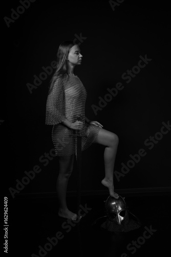 Slender young female knight holding sword and standing with one foot on helmet in black and white