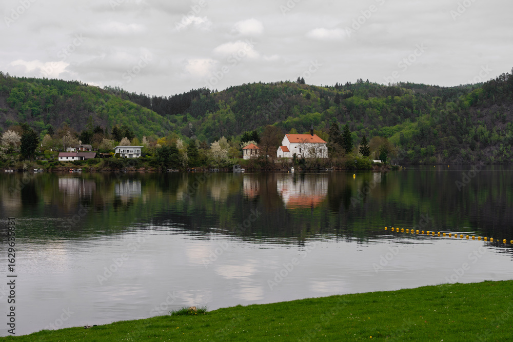Obraz premium Riverside view of white church with red roof and nearby houses reflected in still water. Surrounded by green spring hills and cloudy sky. Peaceful nature scene in Czech countryside. No people visible.
