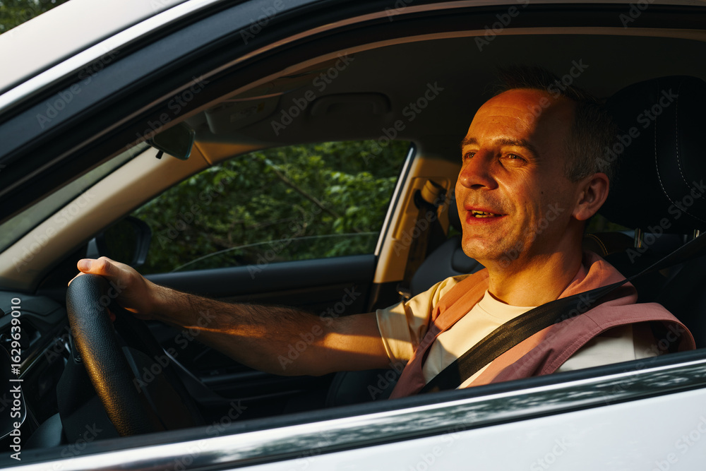 Naklejka premium Middle aged Caucasian man driving car during family road trip, smiling and looking ahead with one hand on steering wheel, sunlight illuminating face, seatbelt fastened