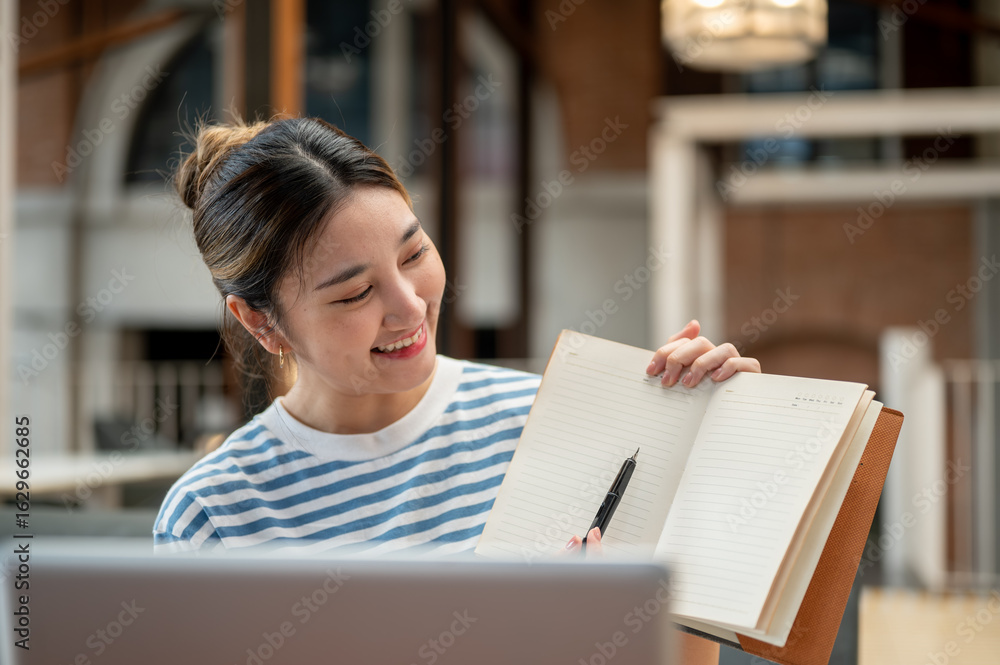 Fototapeta premium Smiling asian young woman holding pen pointing in notebook while sits on sofa across laptop in cafe.