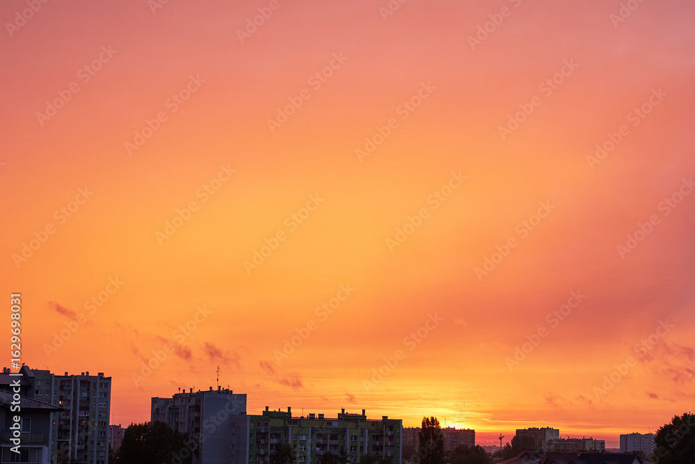 Obraz premium Vibrant orange and pink sky at sunset over urban apartment buildings and city silhouette, showcasing beautiful atmospheric colors and an atmospheric golden hour glow