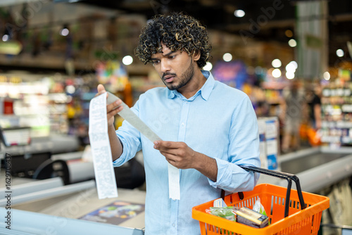Photography A man in a grocery store looks at a long receipt, seemingly surprised by the total cost of his purchase