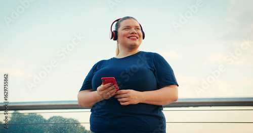 Outdoor Portrait of Body Positive Overweight Woman Using Red Smartphone Phone Outdoors at City in the Morning. Wearing Red Headphones