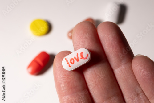 A hand holds a white pill marked with the word love, surrounded by vibrant capsules on a surface. The image symbolizes the complex relationship between addiction, joy, and wellness.
