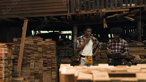 Factory Worker Inspecting Timber And Wood Furniture As Two African American Male Woodworkers Discuss Stock Checking And Quality Inspection In Industrial Workshop