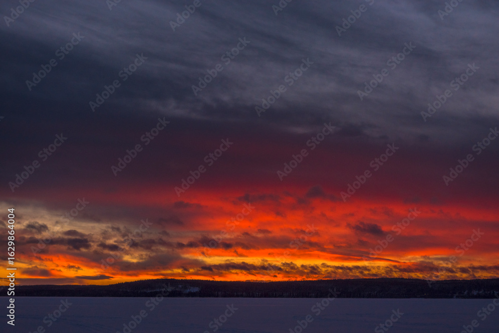 Fototapeta premium A dramatic winter sunset over a frozen lake. Dazzling shades of red, orange and purple break through heavy grey-blue clouds, painting the sky above the horizon in fiery colors. 