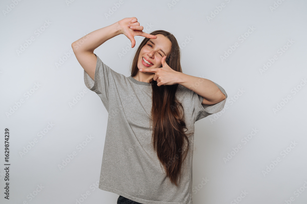Fototapeta premium portrait of young joyful woman playfully frames a scene with her hands, showcasing creativity and a fun on light background