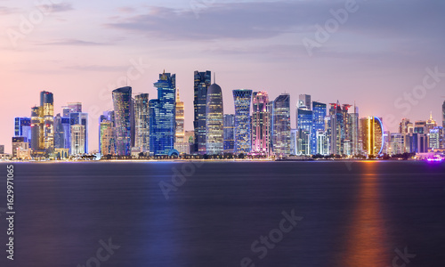 Sunset view of Doha skyline, Qatar, with shades of pink and purple filling the sky. Modern skyscrapers are reflected in the calm waters of the bay, as city lights begin to glow
