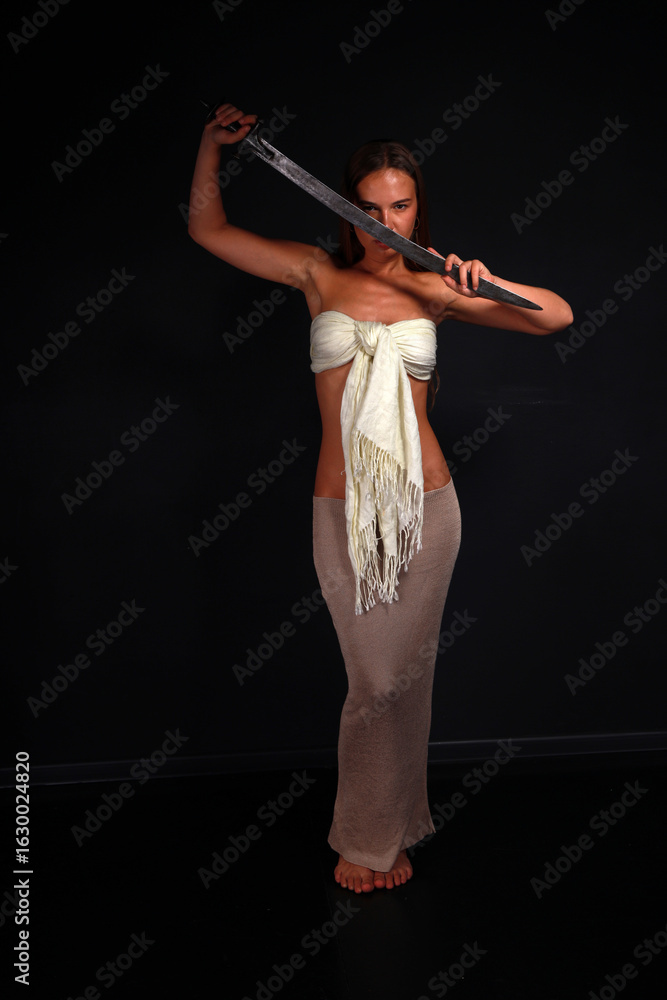 Fototapeta premium Slender young woman in white cloth holding traditional indian sword low key full length portrait