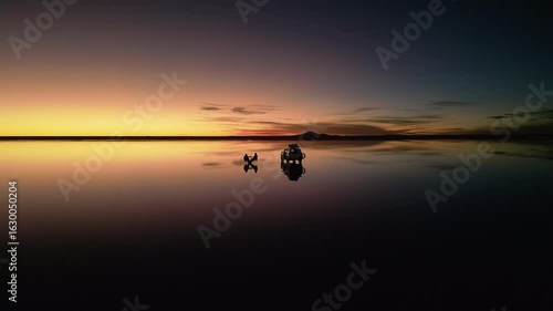 Drone footage circling 4x4 vehicle and two people on chairs at salt flats during sunset. Perfect mirror reflection on water surface. Mountains silhouetted on horizon at dusk.