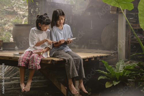 Two Asian children read together on a wooden porch in a rural Thai home, surrounded by warm light, rustic atmosphere, and lush plants. Real Asian countryside lifestyle.