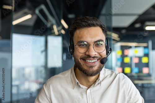 Schilderij op canvas A smiling man wearing a headset, looking directly at the camera in a modern office setting