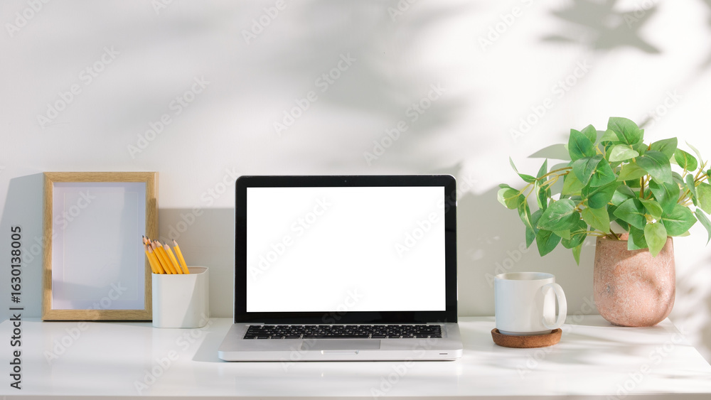 Fototapeta premium Laptop with blank white screen on a white desk, accompanied by coffee mug, and green plant in natural light.