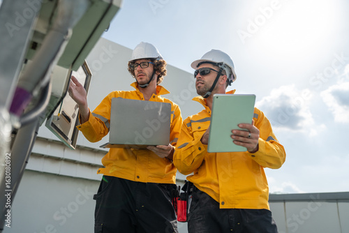 Engineers team in safety helmets and yellow jackets inspecting electrical control cabinet with laptop and tablet on site. Mobile phone Telecommunication Radio antenna tower installed on building