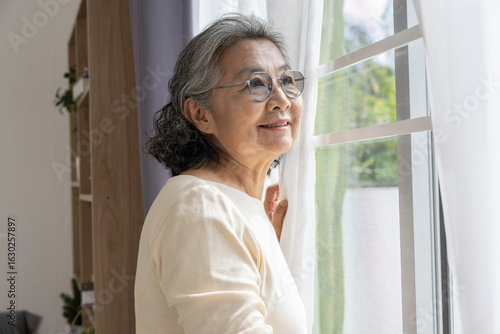 Asian Elderly woman with glasses smiling while looking out window, enjoying natural light and peaceful moment indoors with soft curtains and home decor Free Download Asian Elderly woman with glasses smiling while looking out window, enjoying natural light and peaceful moment indoors with soft curtains and home decor - GFXLoad