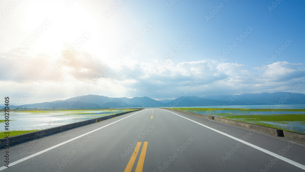 Fototapeta premium Empty asphalt road leading towards distant mountains under a bright blue sky with scattered clouds