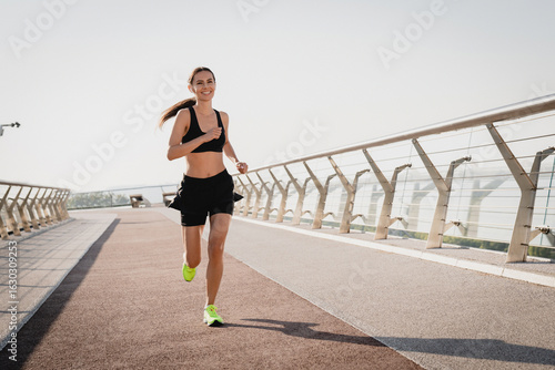 Female runner in activewear burning calories with a cardio workout. Sporty woman jogging running on city park bridge outdoors. Woman exercising with earphones on in the morning. Copy space