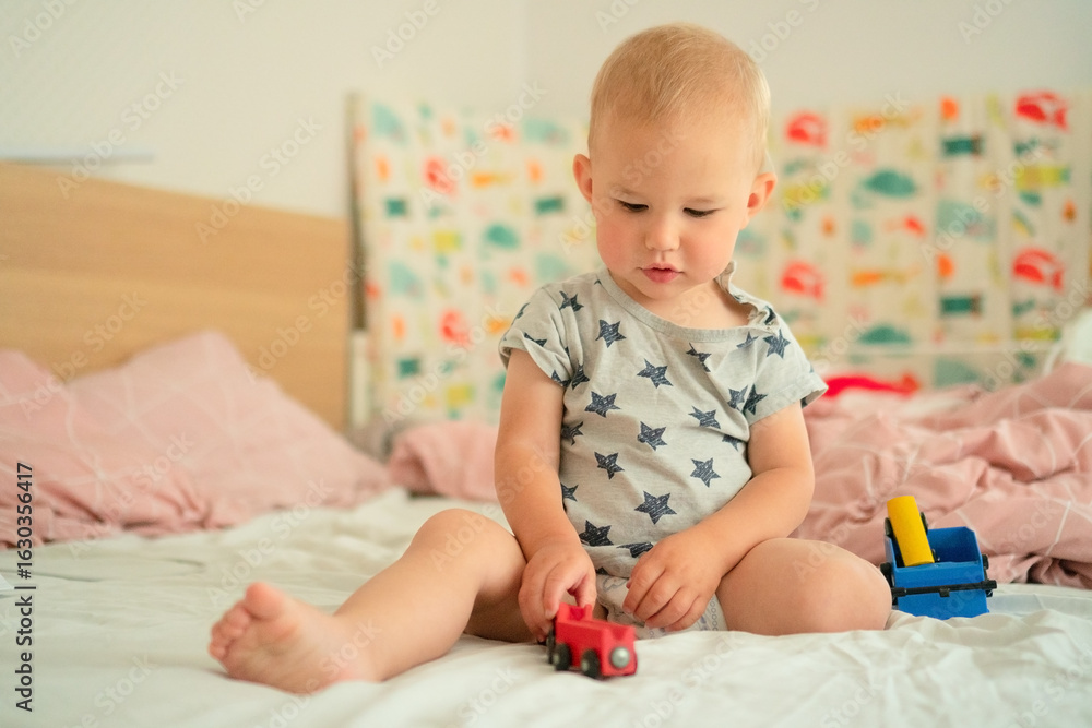 Naklejka premium Cheerful baby in star-patterned romper joyfully plays with a red toy train on a bed. The child exhibits excitement and curiosity, enhancing a playful and warm atmosphere.