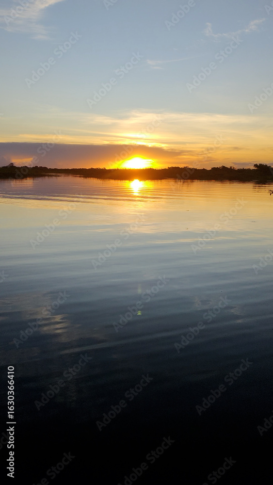 Obraz premium Vertical view of a serene sunset over Cuiabá River in Pantanal wetlands. The golden sun sets on the horizon, casting a vibrant reflection on the calm, rippling water. Nature landscape at twilight.